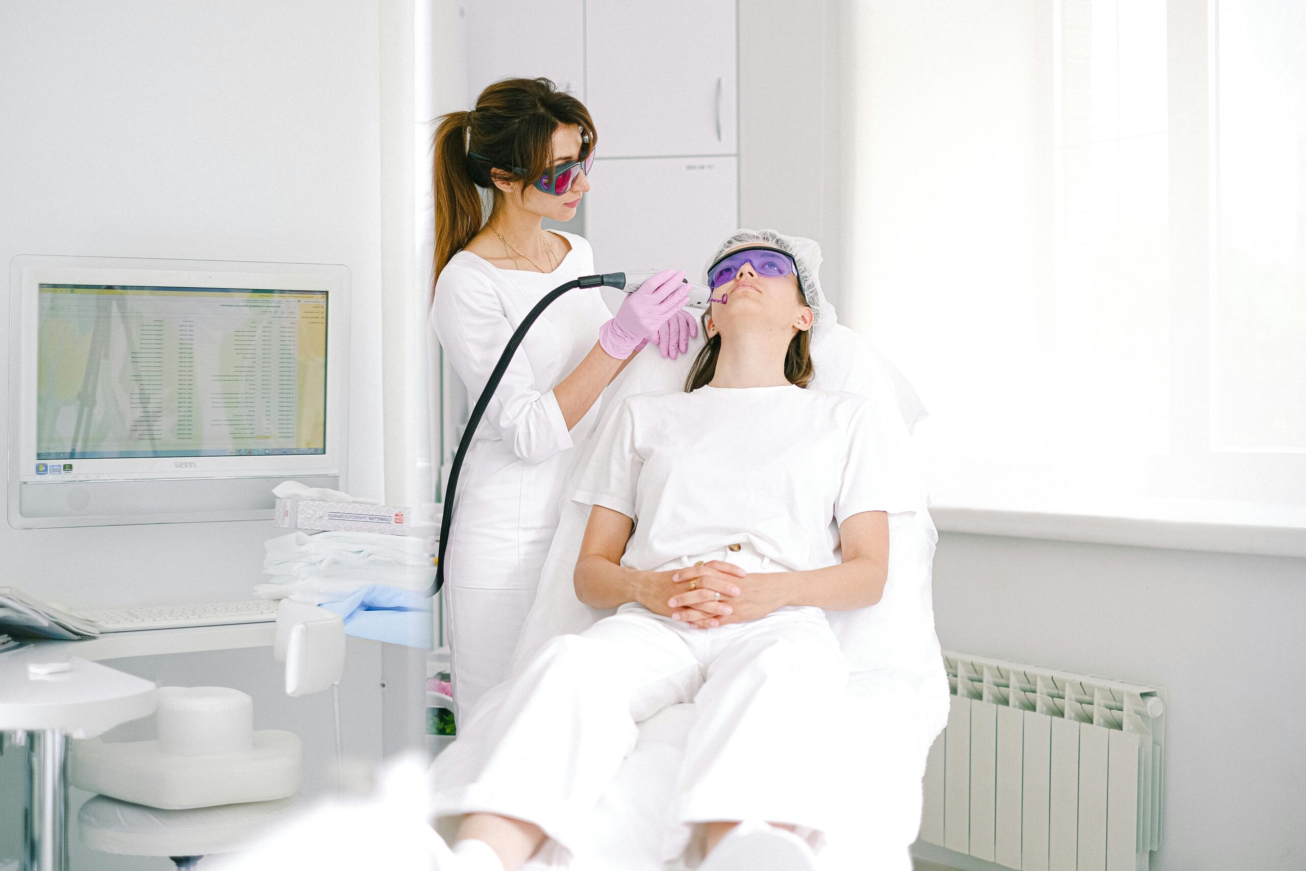 Aesthetician performing laser facial treatment on woman with protective eyewear in a modern clinic.